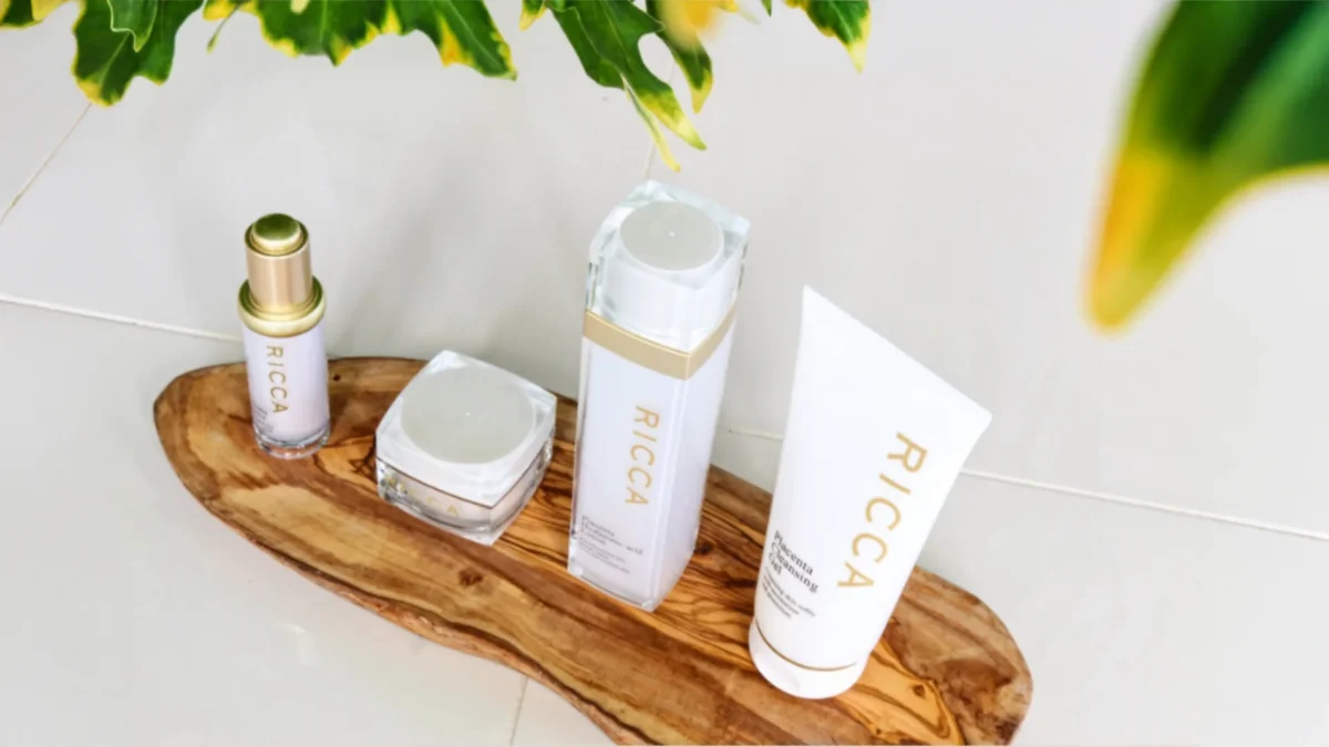 木製のトレーの上に、「RICCA」と書かれたスティックタイプ、クリーム容器、ボトル、チューブタイプの4種類の化粧品が並んでいます。背景は白を基調とした空間で、上部に観葉植物の葉が見えます。