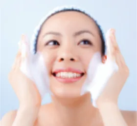 白いヘアバンドを着けた女性が、両手にたっぷりの白い泡をのせて笑顔で頬を洗っているスキンケアのイメージです。