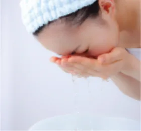白いヘアバンドを着けた女性が洗面ボウルの上で両手に水をため、顔をすすいでいるスキンケアのイメージです。