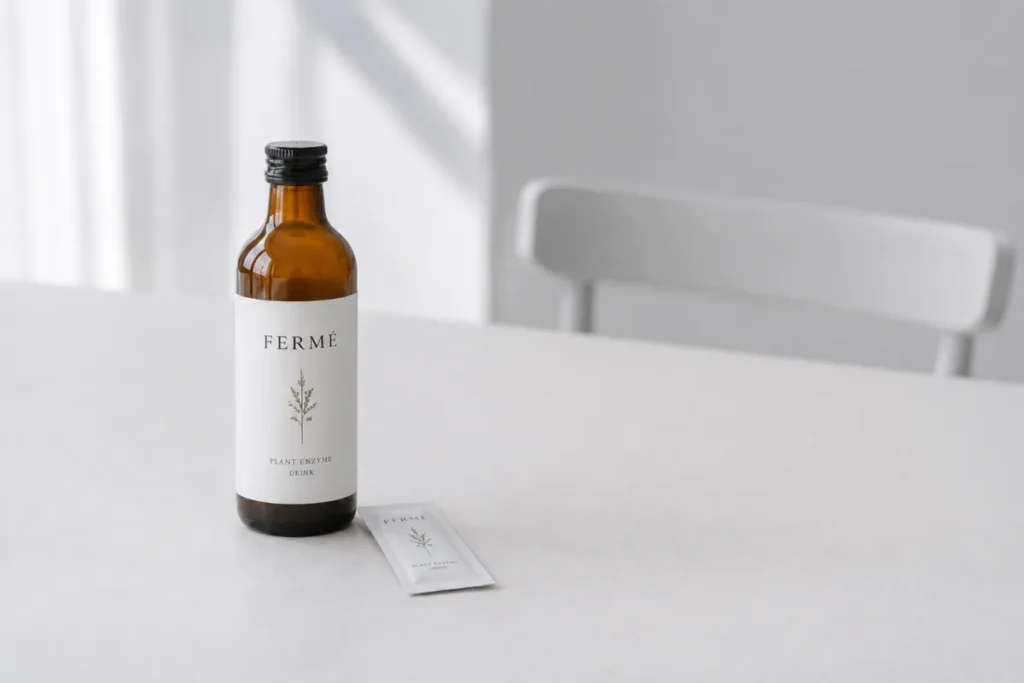 白いテーブルの上に茶色のFERMÉの植物酵素ドリンクボトルと個包装スティックが置かれ、背景に白い椅子と窓から差し込む光が見えます。ボトルと個包装に「FERMÉ PLANT ENZYME DRINK」と書かれています。